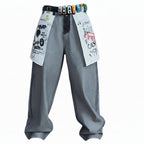 A person wearing loose gray denim pants with graphic patches, a black belt with colorful buckles, and black boots with patterned details, shown against a plain white background.