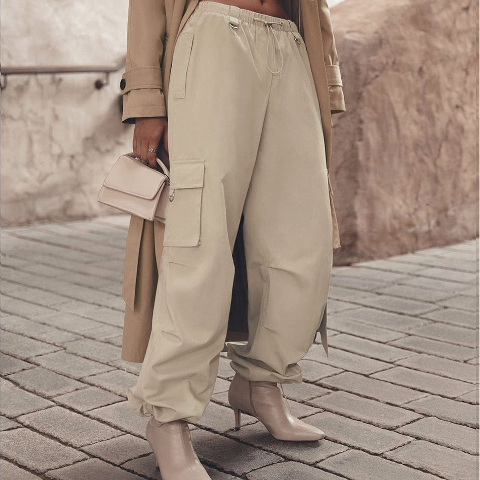 A person wearing loose beige cargo-style pants with an elastic drawstring waist, styled with a beige trench coat, beige handbag, and beige ankle boots on a stone-paved walkway.